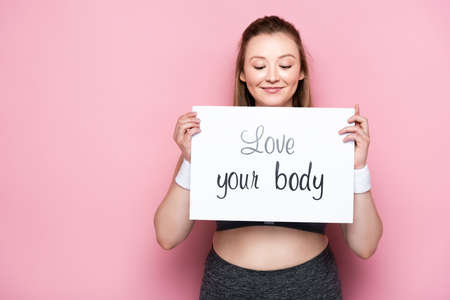 happy overweight girl with closed eyes holding placard with love your body lettering on pinkの写真素材