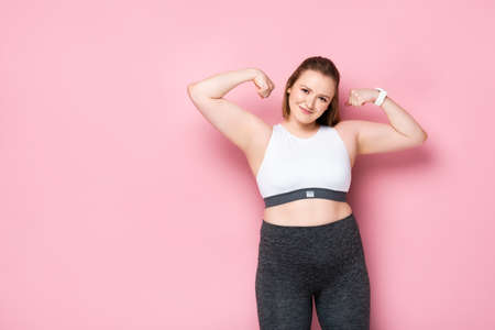 cheerful overweight girl in sportswear demonstrating biceps on pinkの写真素材