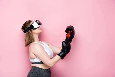 side view of overweight girl in vr headset and boxing gloves looking up on pink