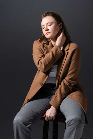 beautiful plus size girl sitting on chair and touching neck while posing with closed eyes isolated on grayの写真素材