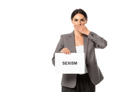 businesswoman covering mouth while holding placard with sexism lettering isolated on white
