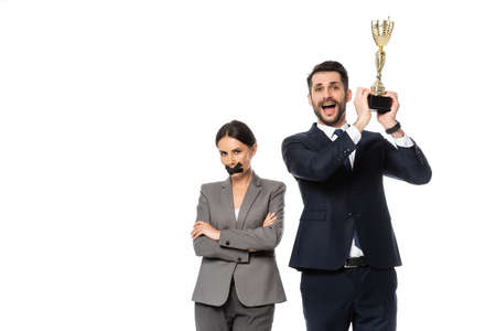 excited businessman holding trophy near businesswoman with duct tape on mouth standing with crossed arms isolated on whiteの写真素材