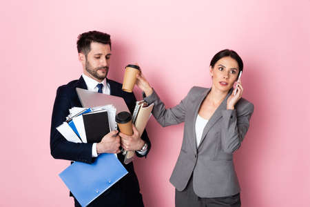 businesswoman talking on smartphone near businessman holding paper cups, laptop, folders and notebooks on pinkの写真素材