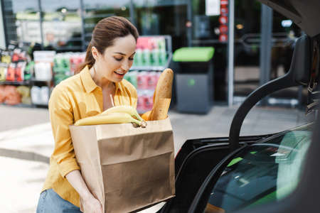 Selective focus of smiling woman holding shopping bag with food near open car trunk on urban street