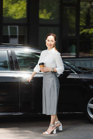 Smiling businesswoman holding paper cup and opening car door on urban streetの写真素材
