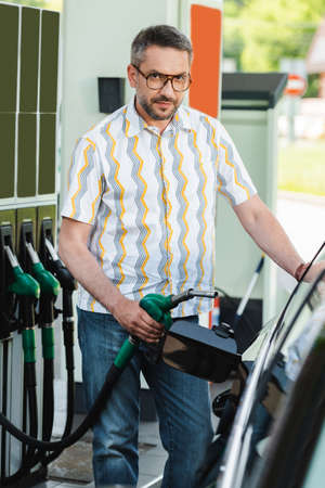 Selective focus of handsome man looking at camera while holding fueling nozzle near car on car refueling stationの写真素材