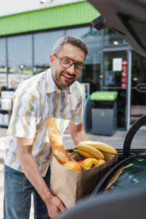 Selective focus of smiling man putting shopping bag with food in open car trunk on urban streetの写真素材