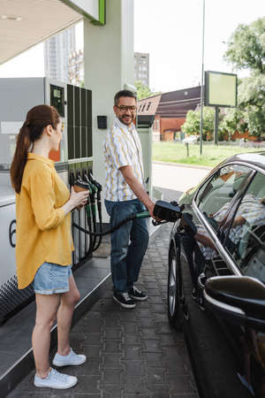 Selective focus of smiling man fueling car near wife with coffee to go on gas station