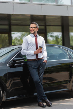 Positive businessman looking away while holding coffee to go near auto on urban streetの写真素材