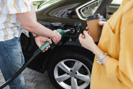 Cropped view of man holding fueling nozzle near gas tank of car near woman with paper cup on gas...