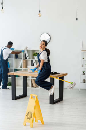 Selective focus of positive cleaner jumping with mop near african american colleague working in...