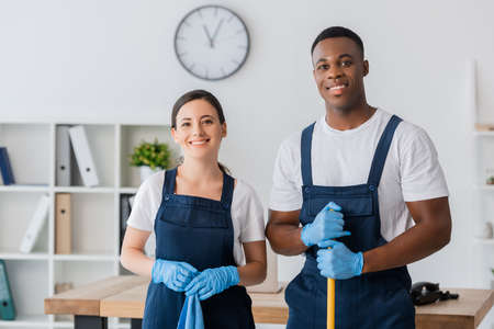 Smiling multiethnic workers of cleaning service holding rag and mop while working in officeの写真素材