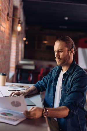 selective focus of bearded businessman looking at charts and graphs near laptop in office