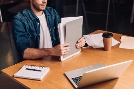 cropped view of businessman holding folder near gadgets and paper cup on desk