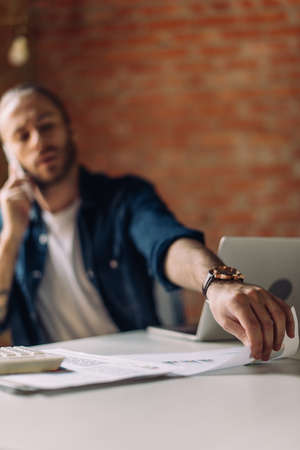 selective focus of businessman talking on smartphone and taking paper near laptop in office