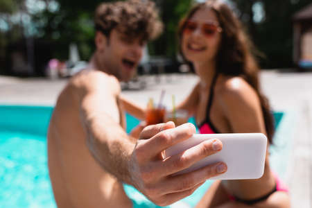selective focus of happy man and woman in sunglasses holding cocktails and taking selfie near...