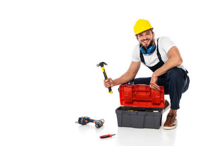 Smiling workman in uniform holding hammer near toolbox and tools on white background