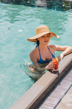 attractive girl in straw hat looking at cocktail in glass while swimming in pool