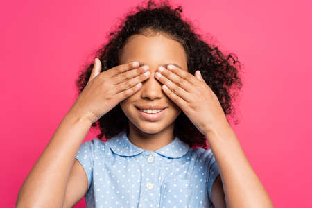smiling cute curly african american kid covering eyes isolated on pink