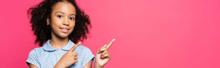smiling cute curly african american kid pointing with fingers aside isolated on pink, panoramic shotの写真素材