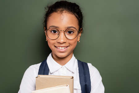 smiling african american schoolgirl with books near empty green chalkboardの写真素材