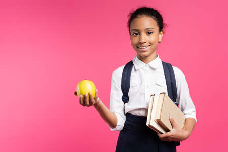 smiling african american schoolgirl with backpack and apple isolated on pinkの写真素材