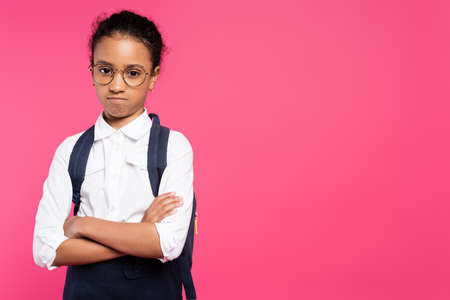 offended african american schoolgirl in glasses with backpack and crossed arms isolated on pinkの写真素材
