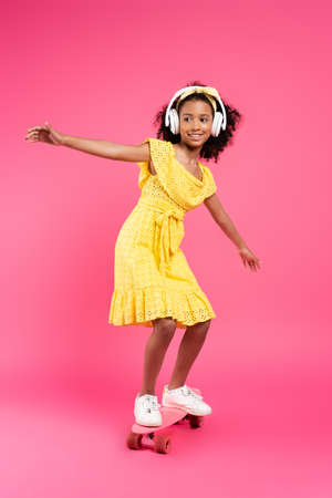 full length view of smiling curly african american child in yellow outfit and headphones riding...