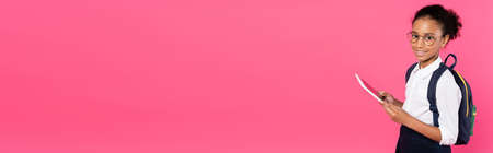 smiling african american schoolgirl in glasses with backpack using digital tablet isolated on pink,...