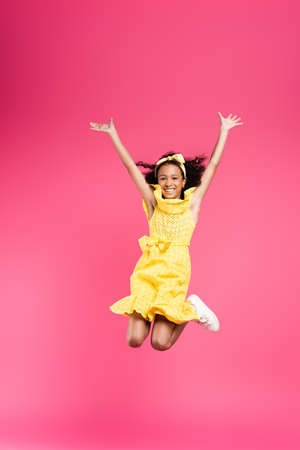full length view of happy curly african american child in yellow outfit jumping with hands in air on...