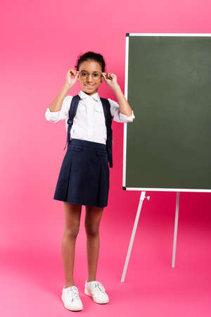 smiling african american schoolgirl with backpack near empty chalkboard on pink backgroundの写真素材