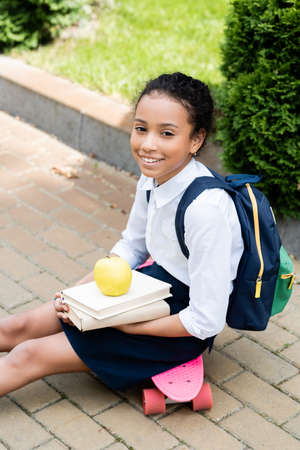 smiling african american schoolgirl with books and apple sitting on penny boardの写真素材