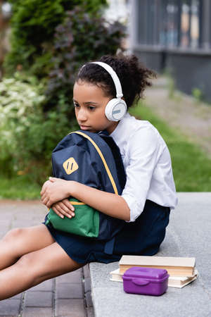 sad african american schoolgirl with backpack and headphones sitting outsideの写真素材