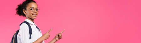 smiling african american schoolgirl in glasses with backpack pointing aside isolated on pink, panoramic shotの写真素材