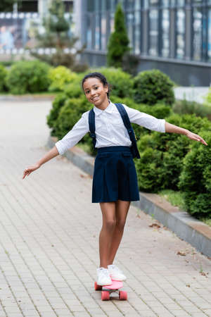 happy african american schoolgirl with backpack riding penny board outdoorsの写真素材