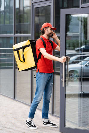 Smiling delivery man talking on smartphone and opening door of building on urban streetの写真素材