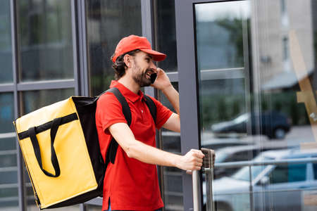 Smiling courier with thermo backpack talking on smartphone while opening door of building on urban streetの写真素材