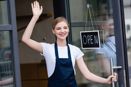 Positive waitress waving hand near signboard with open lettering on door of cafeの写真素材