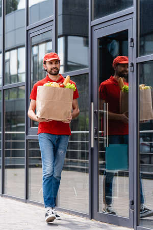 Smiling delivery man holding shopping bag with fresh vegetables while walking on urban streetの写真素材