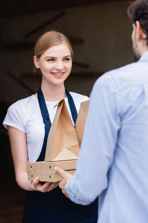 Selective focus of beautiful waitress looking at man and giving pizza boxes and paper bags at entry...