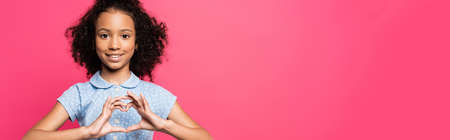 smiling cute curly african american kid showing heart with hands isolated on pink, panoramic shotの写真素材