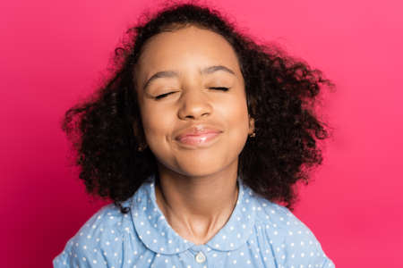 pleased cute curly african american kid with closed eyes isolated on pinkの写真素材