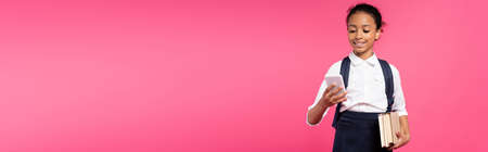 smiling african american schoolgirl with books and smartphone isolated on pink, panoramic shot