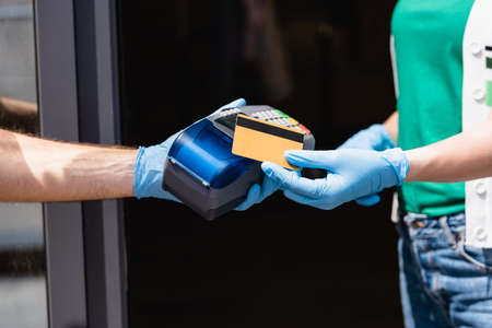 Cropped view of woman in latex gloves holding credit card near courier with payment terminal on...