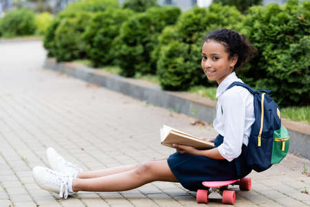 side view of smiling african american schoolgirl reading book while sitting on penny board