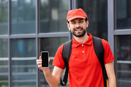 Handsome courier in uniform holding smartphone with blank screen on urban street