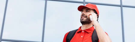 Panoramic crop of smiling courier in uniform talking on smartphone on urban streetの写真素材