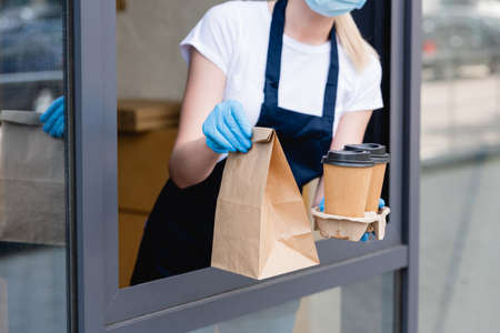 Cropped view of waitress in latex gloves holding paper cups and package near window of cafeの写真素材