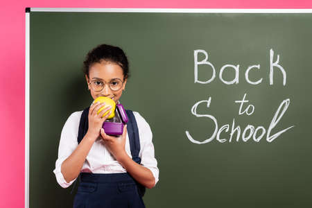 smiling african american schoolgirl in glasses eating apple near back to school inscription on green...