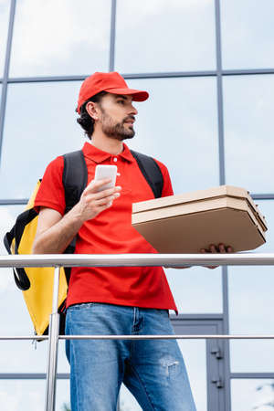 Handsome courier holding pizza boxes and smartphone on urban streetの写真素材